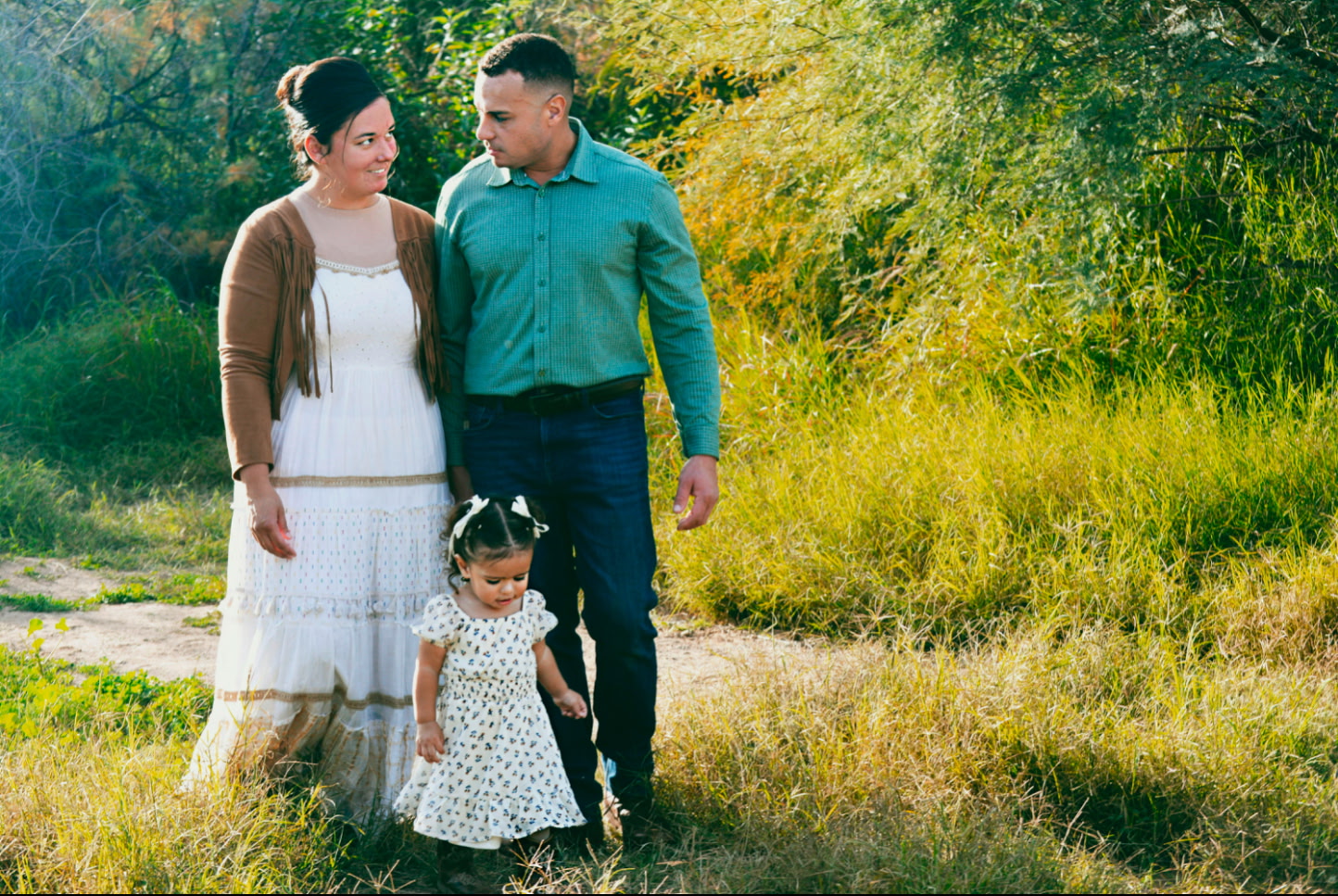 Family of three walking together in a sunlit grassy field; mother in white dress, father in teal shirt, toddler daughter in a floral dress beside them.