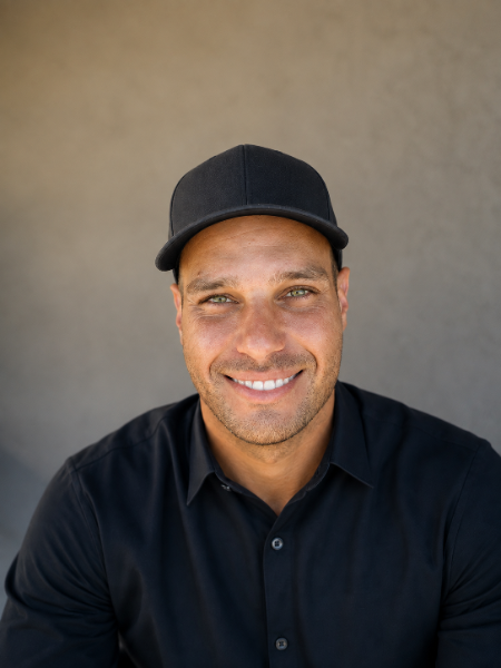 Smiling man in a black cap and black button-up shirt against a gray backdrop.
